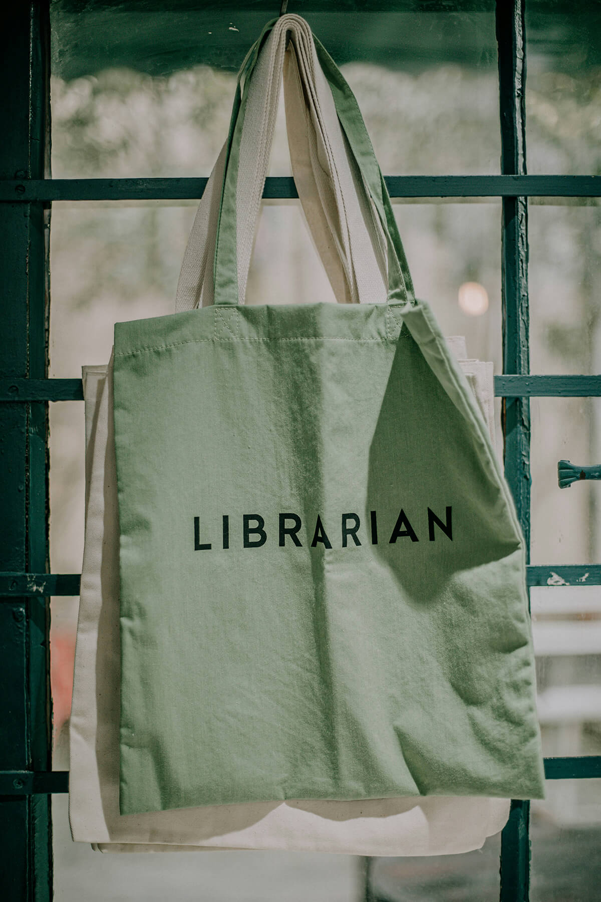 A light green tote bag hanging on a window with the word librarian on it